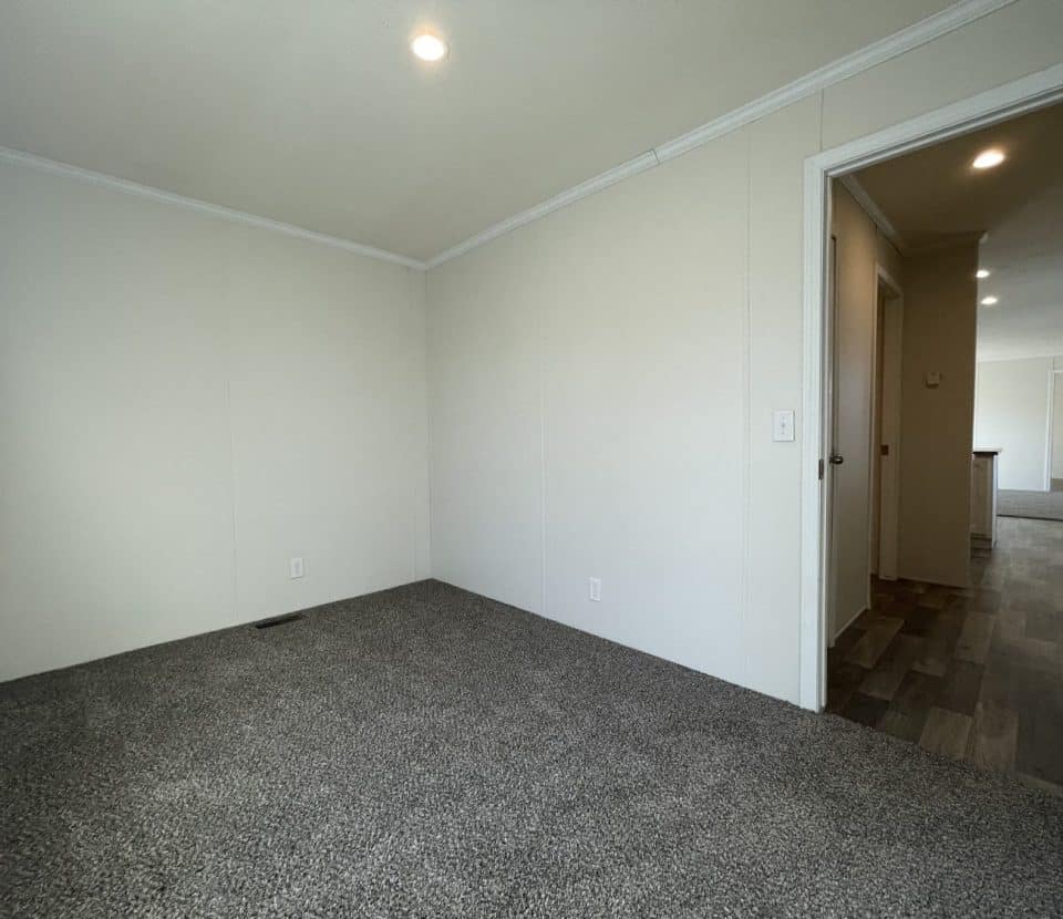 A small, empty room with light-colored walls, gray carpet, and a recessed ceiling light. An open door leads to a hallway with darker wood-style flooring and more rooms visible in the background.