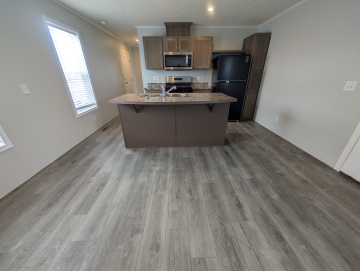 A modern kitchen with wood-look flooring, a center island with a sink, brown cabinets, a black refrigerator, and stainless steel microwave and stove. There is natural light coming in from a window on the left.