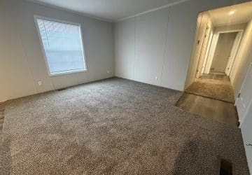 Empty room with light gray carpet, white walls, and a single window with closed blinds. The room connects to a hallway with wood flooring and additional doors visible down the hall.