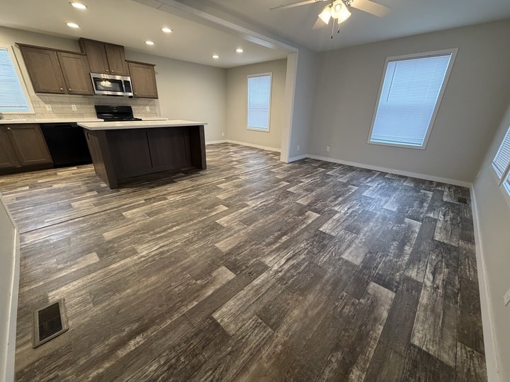Open-plan kitchen and living area with dark wood cabinets, island, and stainless steel appliances. The room features gray wood-look flooring, recessed lighting, and large windows with white blinds.