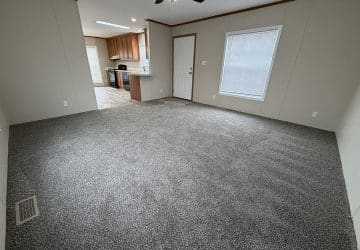 A spacious living room with gray carpet, beige walls, a ceiling fan with a light, and an open view into a kitchen with wooden cabinets and appliances. Theres a window and a door on one wall.