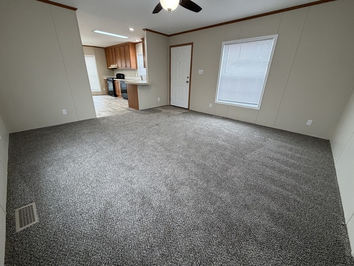 A spacious living room with gray carpet, beige walls, a ceiling fan with a light, and an open view into a kitchen with wooden cabinets and appliances. Theres a window and a door on one wall.