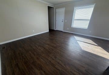 A bright, empty room with beige walls, dark wood flooring, a white door, and a large window letting in sunlight.