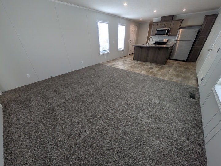 Open living space with gray carpet leading to a tiled kitchen area. The kitchen has dark wood cabinets, stainless steel appliances, and two large windows letting in natural light. The room appears empty and freshly cleaned.