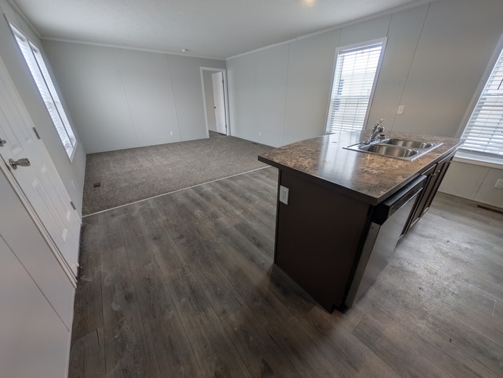 Open-plan room with wood-look flooring and carpeted living area, an island with a sink in the kitchen, large windows with blinds, and white walls with minimal decor.