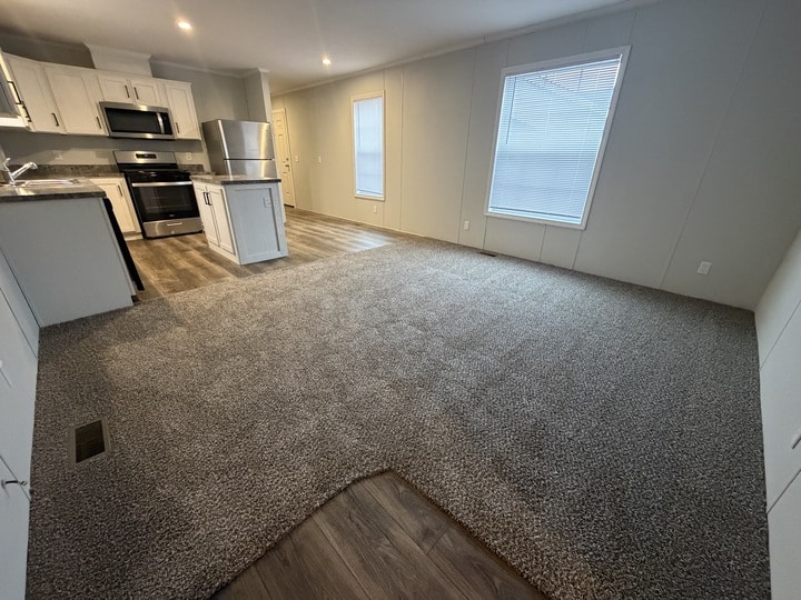 Open living space with gray carpet transitioning to wood flooring. Kitchen area features white cabinets, stainless steel appliances, and island. Large window with blinds lets in natural light. Walls are light colored.