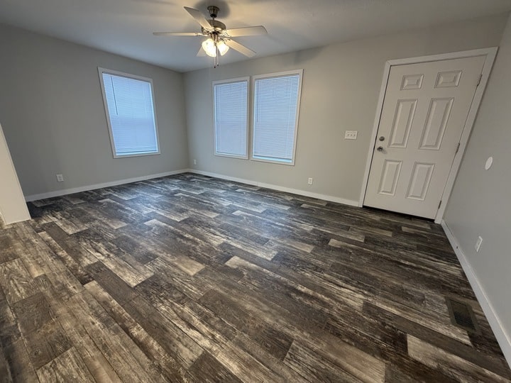 Empty room with dark wood-patterned flooring, light gray walls, two large windows with blinds, a white ceiling fan with lights, and a white front door. The space appears clean and modern.