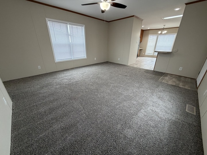A spacious, empty living room with gray carpet, beige walls, a ceiling fan with lights, and a large window. The room opens into a tiled kitchen and dining area with overhead lighting.