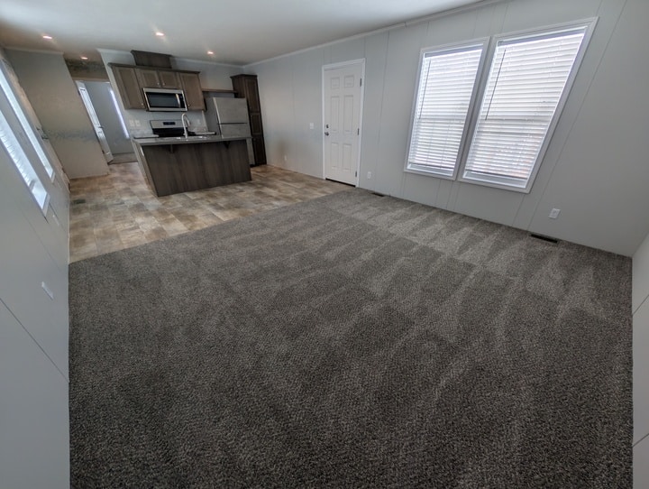 Spacious living area with clean, patterned carpet, large windows with white blinds, and an open view of a modern kitchen with wood cabinets and stainless steel appliances.