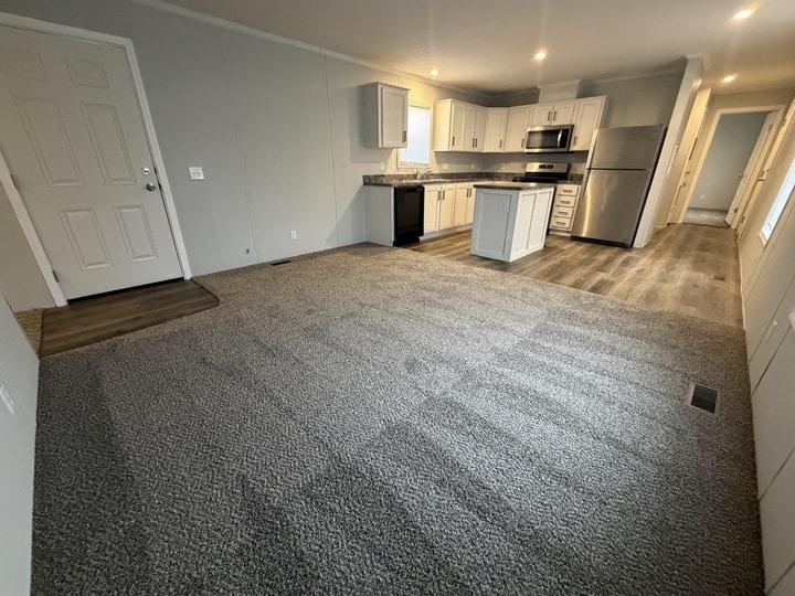 Open-plan room with carpeted living area and wood-look floors in the kitchen. The kitchen features white cabinets, stainless steel appliances, and an island. A hallway and entry door are visible. The space is empty and well-lit.