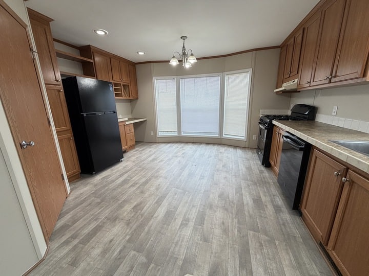 Spacious kitchen with wood cabinets, black appliances including a refrigerator, dishwasher, and stove, light wood flooring, large windows with blinds, and overhead lighting.