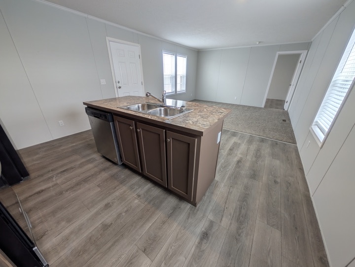 Open kitchen with a central island featuring a double sink and dishwasher, brown cabinets, wood-style flooring, and an adjacent carpeted living area with large windows and white walls.
