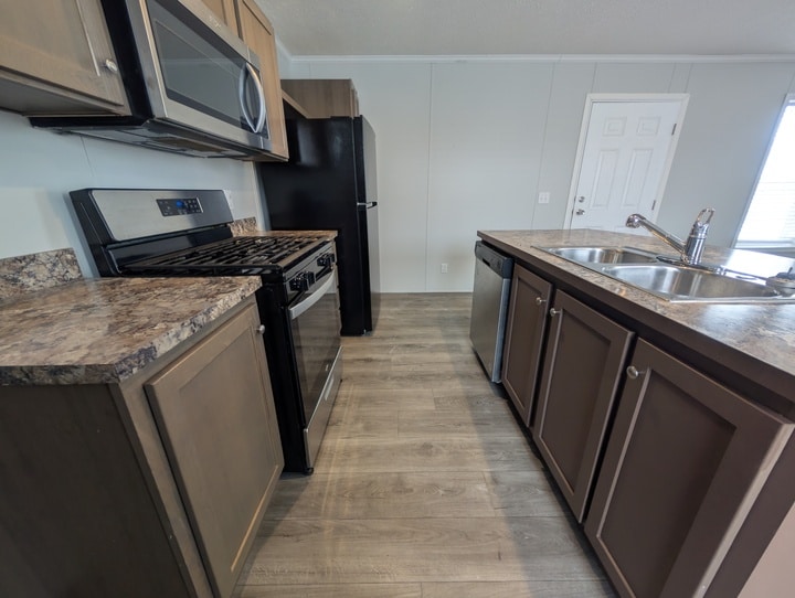 Modern kitchen with wood cabinets, marble-look countertops, stainless steel sink, dishwasher, gas stove, microwave, black refrigerator, and light wood flooring. A white door and window are visible in the background.