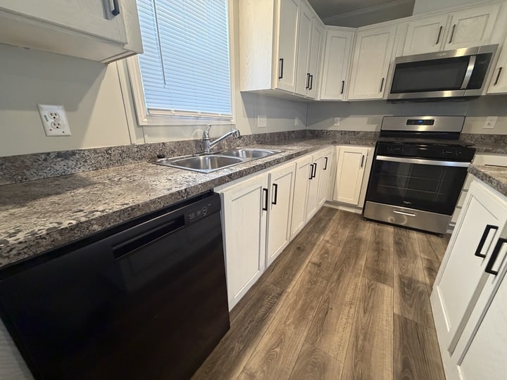 Modern kitchen with white cabinets, granite countertops, stainless steel sink, black dishwasher, and black oven with a microwave above. The floor is wood-patterned laminate, and a window is above the sink.