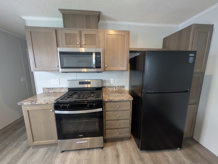 A modern kitchen with wood cabinets, a stainless steel gas stove and oven, a microwave above the stove, granite-pattern countertops, and a large black refrigerator. The floor has a wood-like finish.