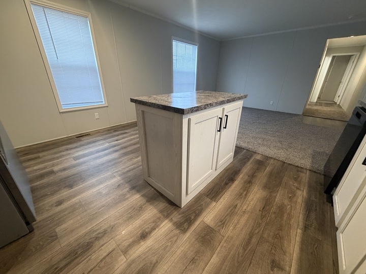 A modern kitchen with wood-look flooring, a small island with a marble countertop and cabinet storage, and an open view into a carpeted living area and hallway with several doors.
