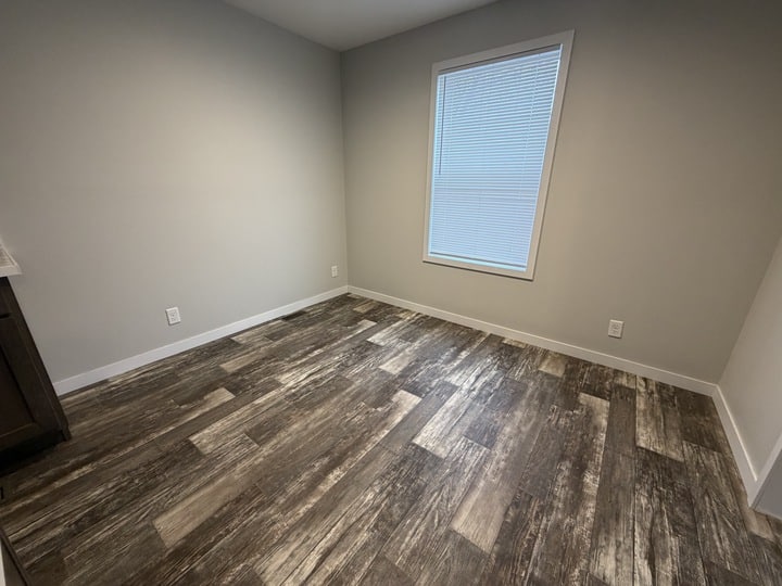 Empty room with light gray walls, a large window with closed blinds, wood-look flooring in shades of brown and gray, and several electrical outlets along the walls.