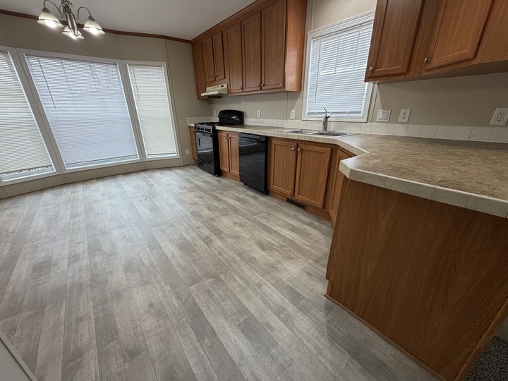 A spacious kitchen with light wood cabinets, beige countertops, black appliances, and gray wood-look flooring. Large windows with blinds let in natural light, and a small chandelier hangs from the ceiling.
