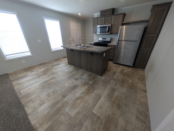 Modern kitchen with wood-style flooring, an island with a sink, stainless steel refrigerator, stove, and microwave. Two large windows and a door provide natural light. The space has neutral-toned walls and cabinets.