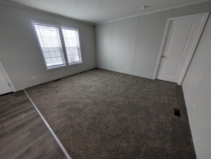 A mostly empty room with light gray walls, a window with white blinds, a closed white door, and brown carpet. Part of the floor near the entrance is covered with gray wood-like flooring.