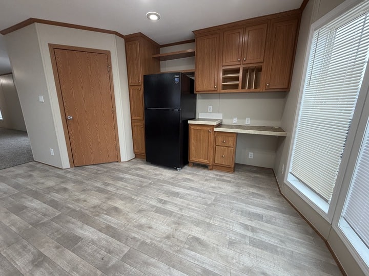 A kitchen area with wood cabinets, a black refrigerator, open shelves, a small wine rack, and a desk-like counter. The floor is light wood, and large windows let in natural light. A closed wooden door is on the left.