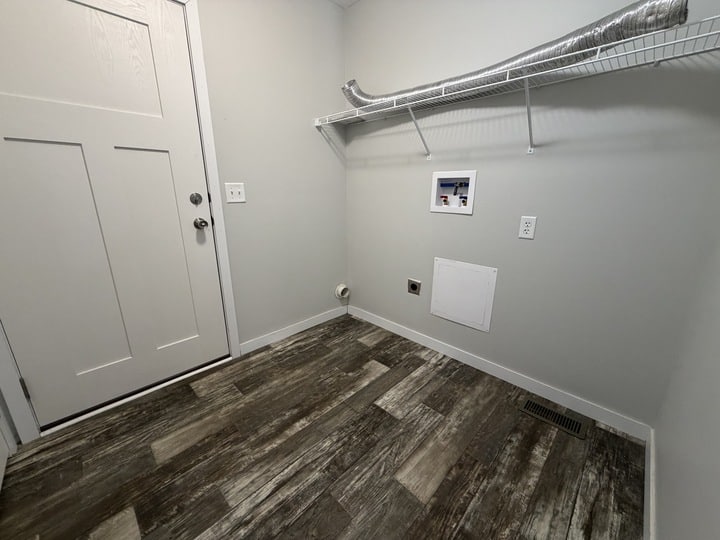 A laundry room with gray walls, wood-look flooring, a white door, a wire shelf, and hookups for a washer and dryer. An exposed duct runs along the shelf on the right wall.