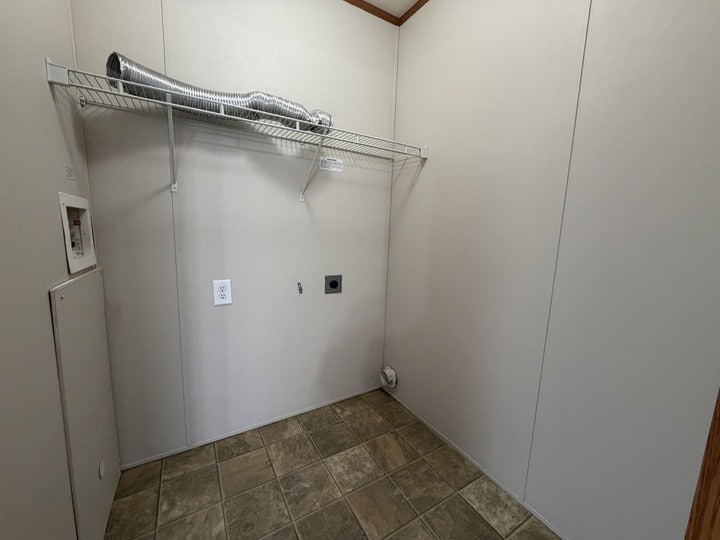 Empty laundry room with beige walls, tile floor, a wire shelf holding a flexible dryer vent hose, and connections for a washer and dryer on the wall. A small utility panel is visible on the left.