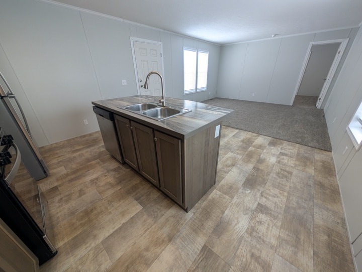 Modern kitchen with wood-look flooring, a center island with a sink and dishwasher, and an open view into a carpeted living area and adjacent rooms; large windows provide natural light.