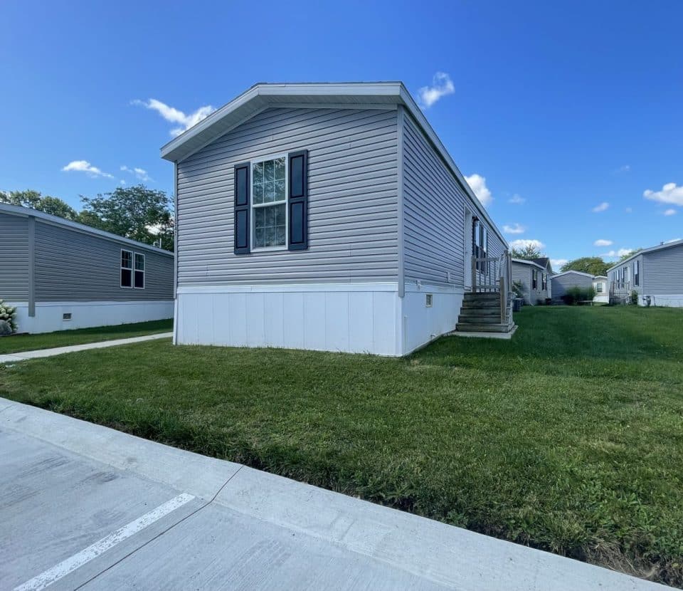 A light gray manufactured home with white trim and black shutters sits on a green lawn with a concrete sidewalk and driveway under a blue sky with scattered clouds. Similar homes are visible in the background.