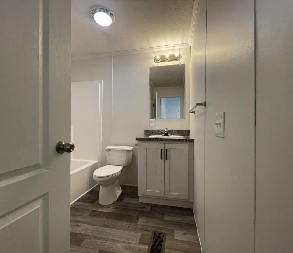 A small, clean bathroom with gray wood-look flooring, a white toilet, a white vanity with a granite countertop, a rectangular mirror above the sink, and a bathtub partially visible on the left.