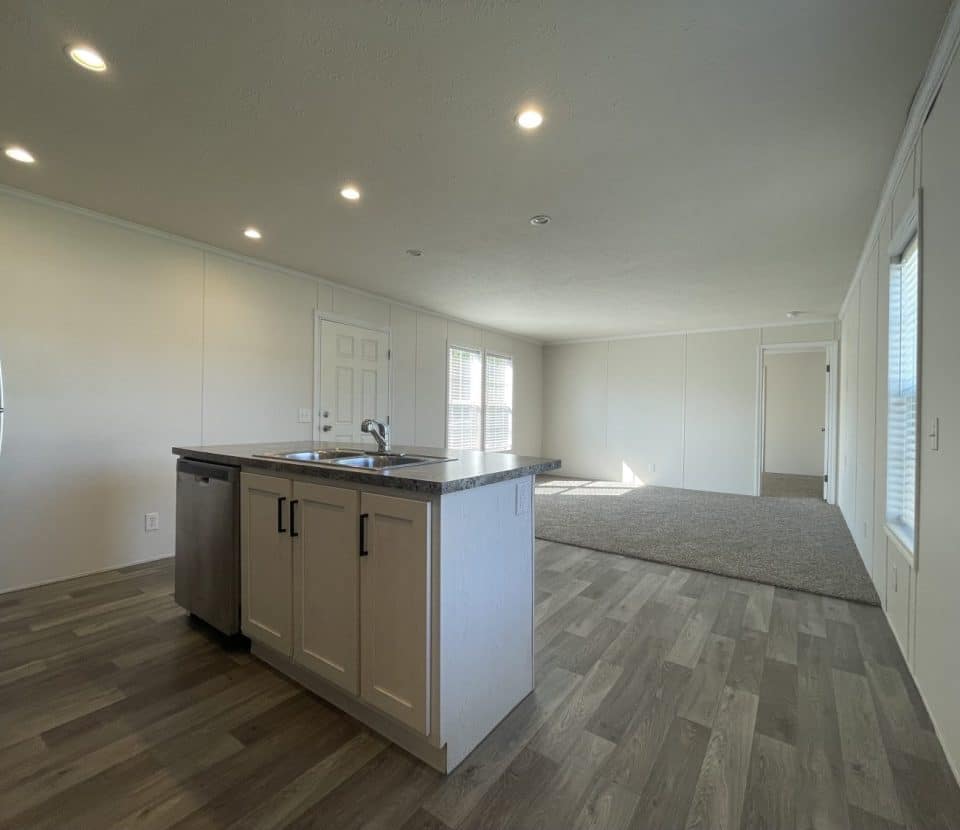 Modern open-concept kitchen and living area with wood-look flooring, central island with sink, stainless steel appliances, white walls, and large windows letting in natural light.
