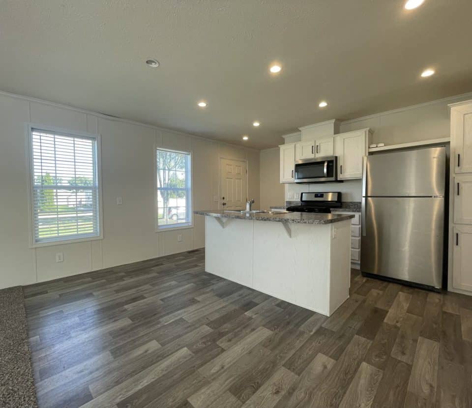 Modern kitchen with stainless steel appliances, white cabinets, a center island, and wood-style flooring. Large windows let in natural light, and recessed lights are on the ceiling. The front door is visible in the background.