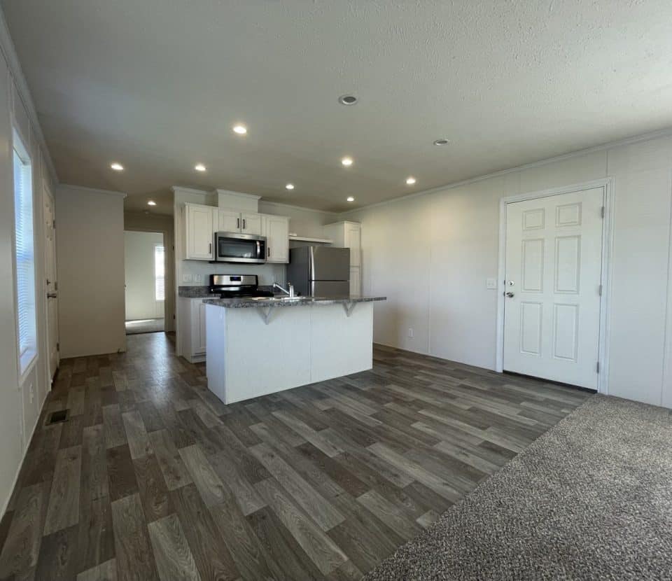 A modern, unfurnished open-plan kitchen and living area with wood-patterned flooring, recessed lighting, white cabinets, stainless steel appliances, and a white door. Light streams in from multiple windows.