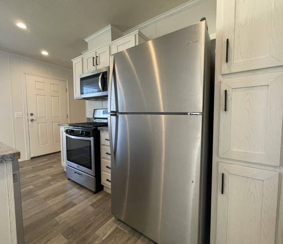 Modern kitchen with a stainless steel refrigerator, gas stove, microwave, white cabinets, and wood-look flooring. The kitchen is well-lit with recessed ceiling lights and a door visible in the background.