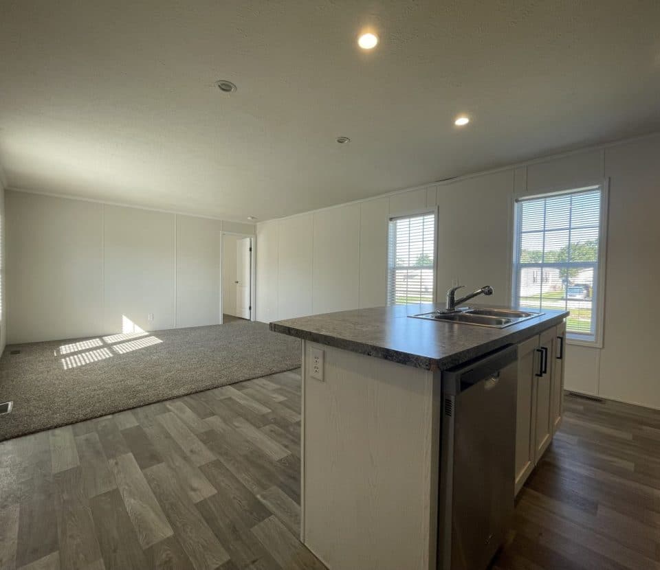 Interior of a modern, unfurnished home featuring a kitchen island with a sink and dishwasher, wood-look flooring, and an open living area with carpet, large windows, and white walls.