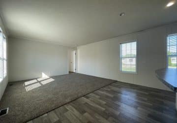 A bright, empty room with white walls, large windows, gray wood-look flooring, and a section of carpeted area. Sunlight streams in, and a door is visible at the far end of the room.
