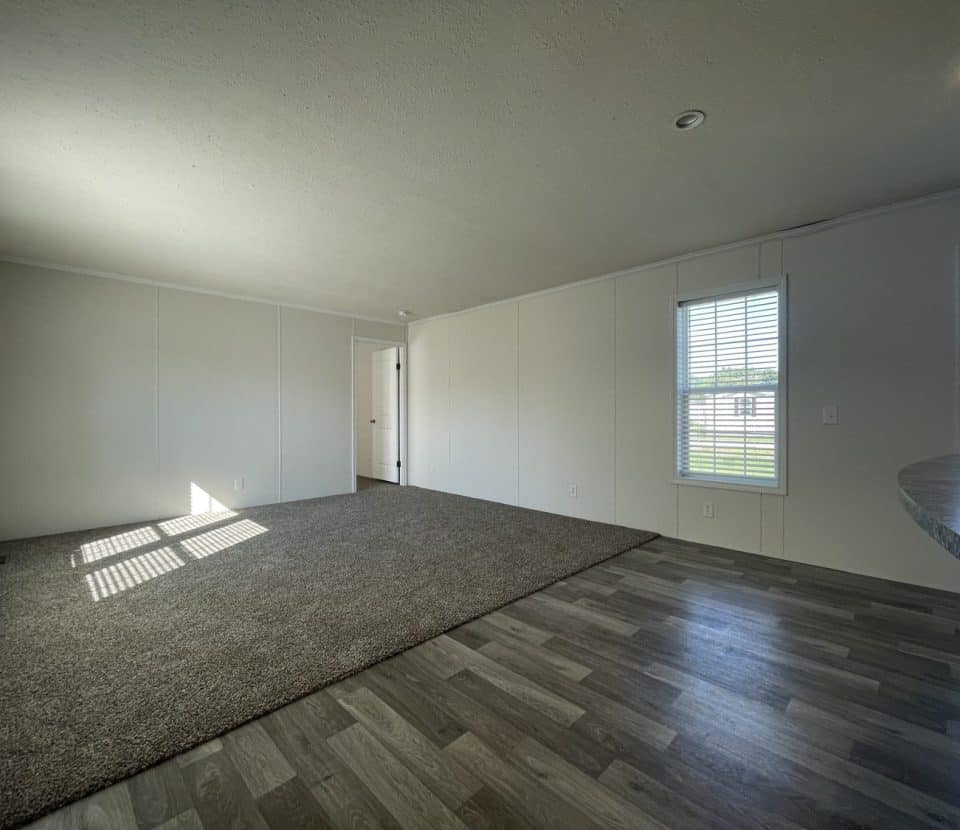 A bright, empty room with white walls, large windows, gray wood-look flooring, and a section of carpeted area. Sunlight streams in, and a door is visible at the far end of the room.