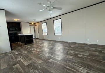 Spacious open-plan living area with wood-look flooring, neutral walls, ceiling fan, and large windows. The modern kitchen features dark cabinets and appliances, with an island and overhead lighting.