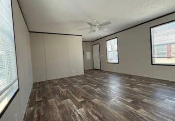 A spacious, empty room with wood-look flooring, light beige walls, three windows, a white ceiling fan, and a white door; sunlight streams through the windows, creating a bright atmosphere.