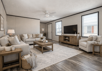 A modern living room with light-colored sofas, a wooden coffee table, an armchair, and a flat-screen TV on a wooden stand. The room has large windows, neutral decor, houseplants, and wood-look flooring.
