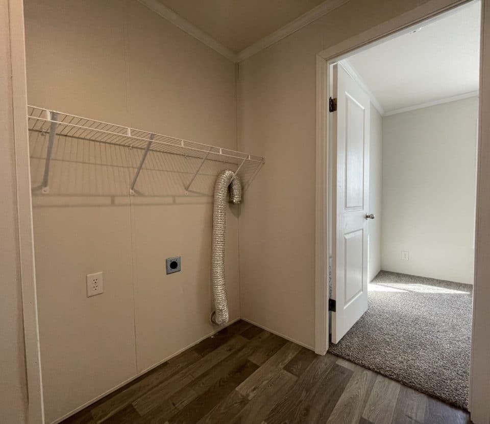 Empty laundry room with beige walls, a wire shelf, dryer vent hose, electrical outlets, and a tile floor. An open door leads to a carpeted room filled with natural light.