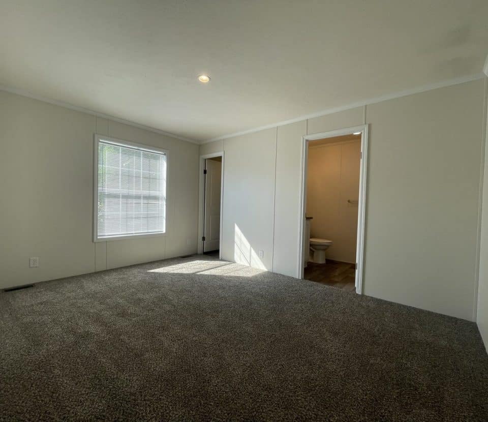 A bright, empty room with beige walls, a single window with blinds, gray carpet, and two open doorways—one leading to a small bathroom with a toilet visible.