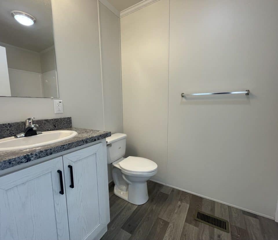 A clean bathroom with a white sink and cabinet, a granite countertop, a toilet, a towel bar on a plain wall, and gray wood-patterned flooring. A mirror and part of a bathtub are also visible.