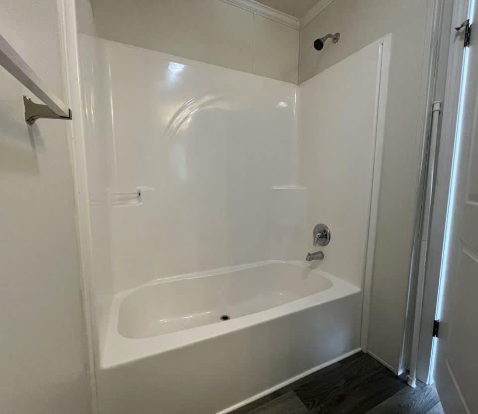 White bathtub and shower combo with a built-in ledge, chrome faucet, and showerhead. The surrounding walls are plain, with a towel bar on the left and a door partially open on the right. The floor has dark wood-like tiles.