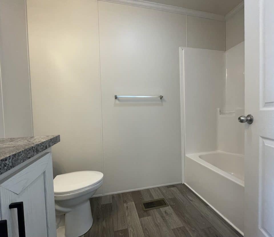 A simple bathroom with light-colored walls, a toilet, a towel bar, a bathtub with a shower, a white door, a vanity with a granite countertop, and wood-style flooring.