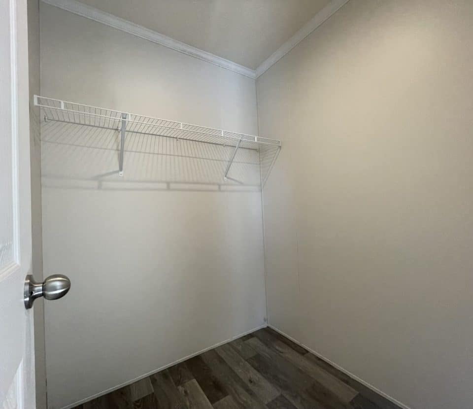 A small empty closet with white walls, a wire shelf on the left wall, a metallic door handle, and wood-patterned laminate flooring.