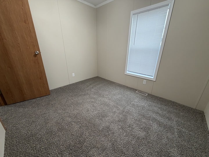 A small, empty room with beige walls, a closed wooden door, gray carpet, and a window with closed white blinds letting in light. An electrical outlet and a floor vent are visible near the window.