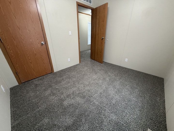 A small, empty room with beige walls, gray carpet, and two wooden doors—one closed and one open, revealing a hallway and part of a window in the adjacent room.