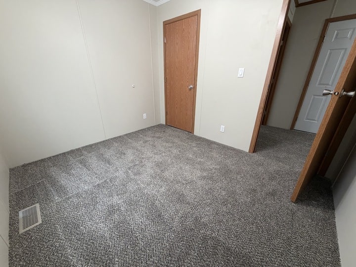 Empty room with beige walls, gray carpet, and three wooden doors; one door is partially open leading to another carpeted space, while another closed door is on the left wall. A vent is visible on the floor.
