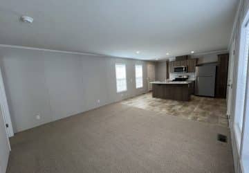 Open-plan room with beige carpet, neutral walls, and a kitchen area with tiled flooring, stainless steel appliances, and an island. Three large windows let in natural light. Room is unfurnished.
